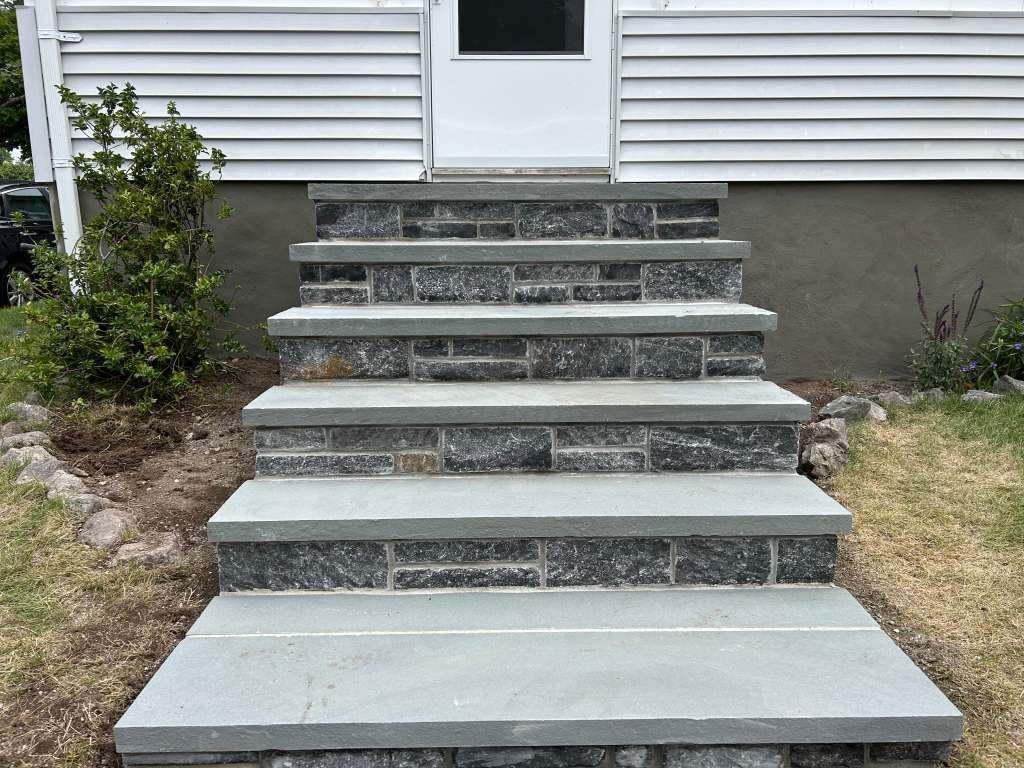 Concrete front steps leading to a white house door, with gray siding and small shrubs on both sides.