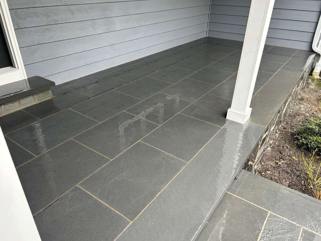 Covered porch with gray tile flooring, white siding, and a small garden bed on the right.
