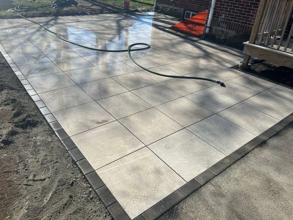 Patio with light gray pavers, a coiled black hose, and a small wet area in sunlight.