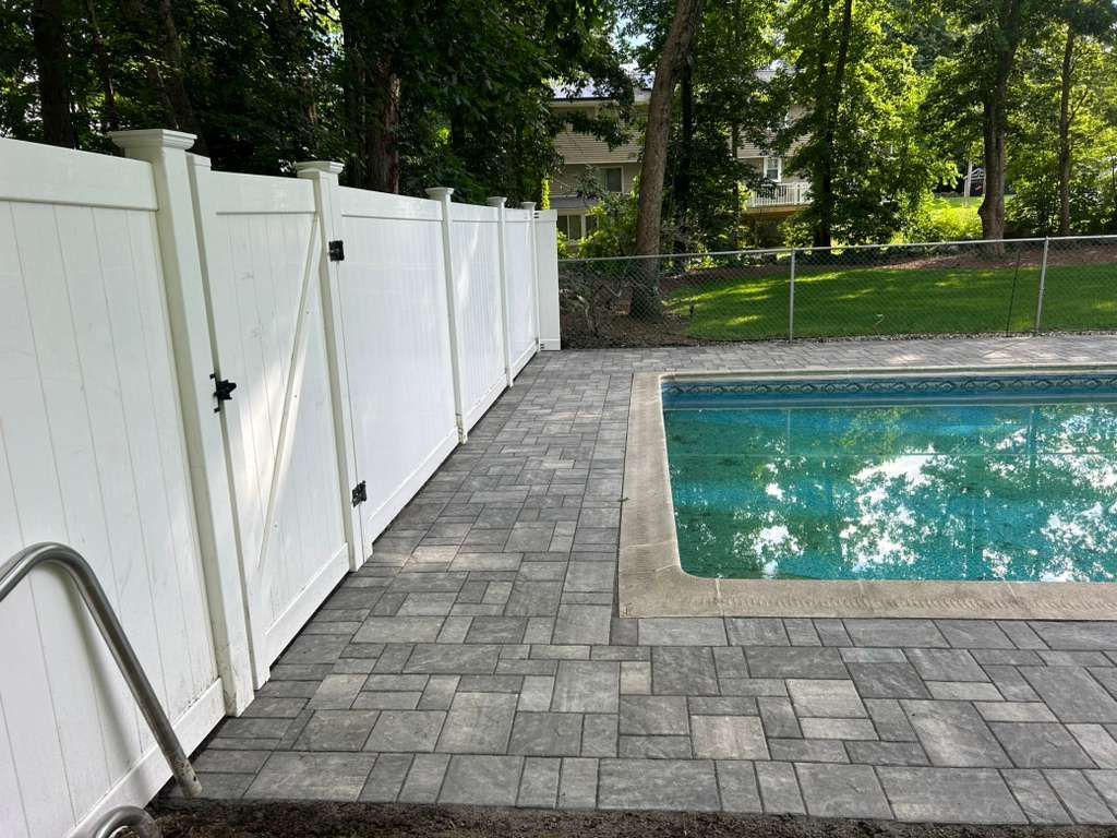 White fence beside a stone patio and swimming pool in a sunny backyard.