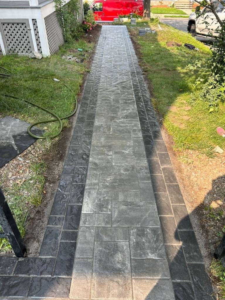 Narrow gray paver walkway leading from a yard to a red gate, bordered by grass and plants.
