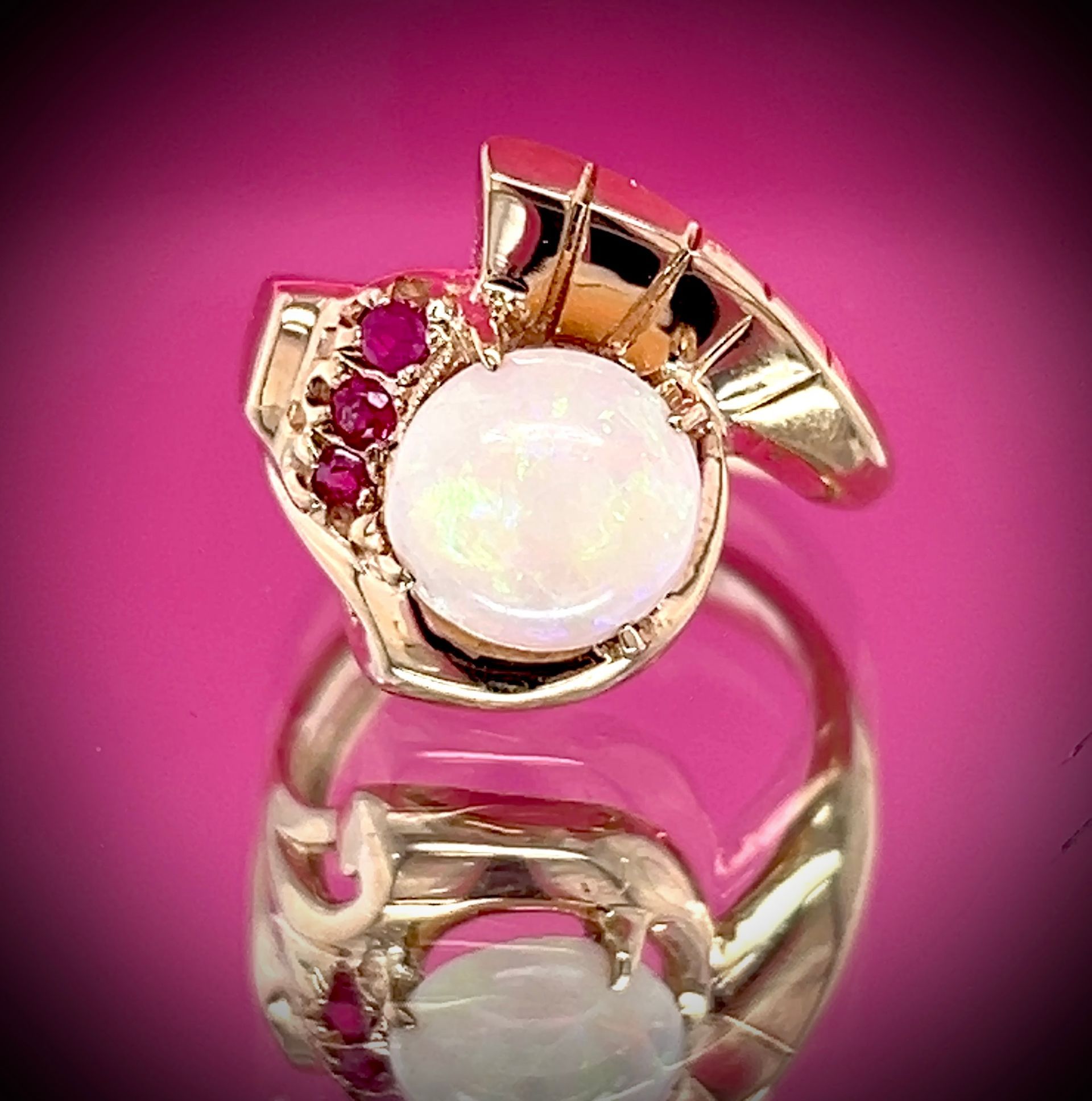 Gold ring with a large white opal and surrounding red stones; reflected in pink background.