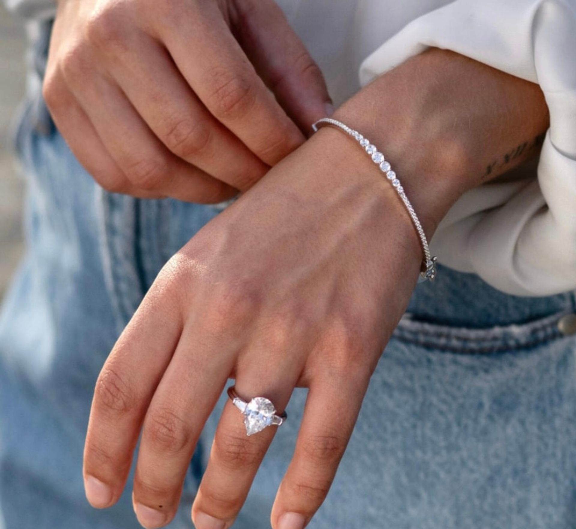 Person's hand with a pear-shaped diamond ring and a delicate bracelet. Leaning toward the bracelet.