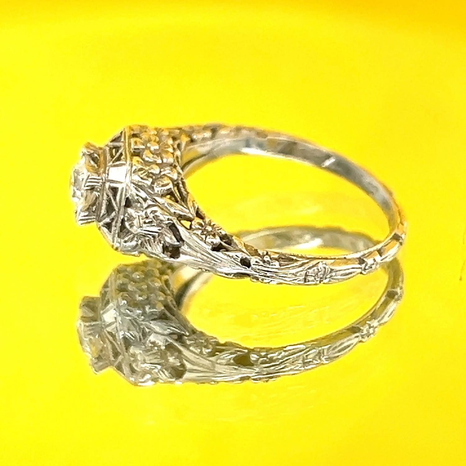 Ornate, white gold ring with a diamond center stone, resting on a yellow surface with its reflection visible.