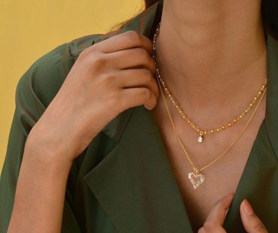 Woman wearing gold layered necklaces, holding the top of her olive-green jacket, with a yellow background.