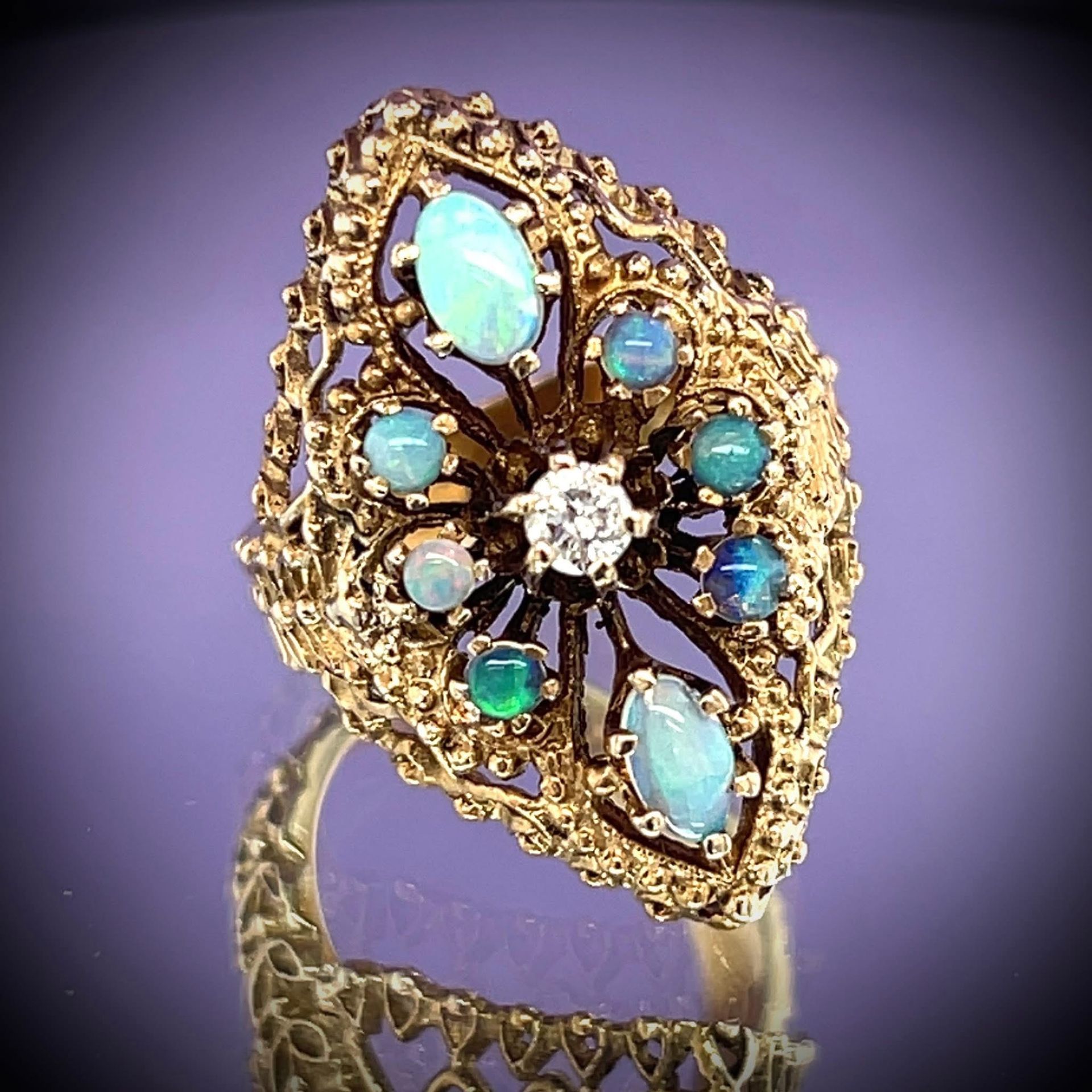 Gold ring with opals and a central diamond, ornate filigree design, reflecting on a purple surface.