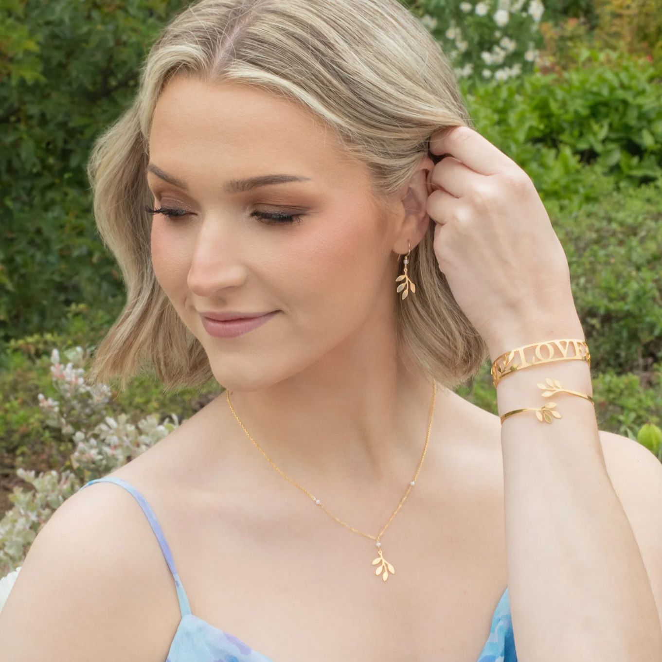 Woman wearing gold jewelry, looking down with a slight smile. Outdoors setting with greenery in the background.