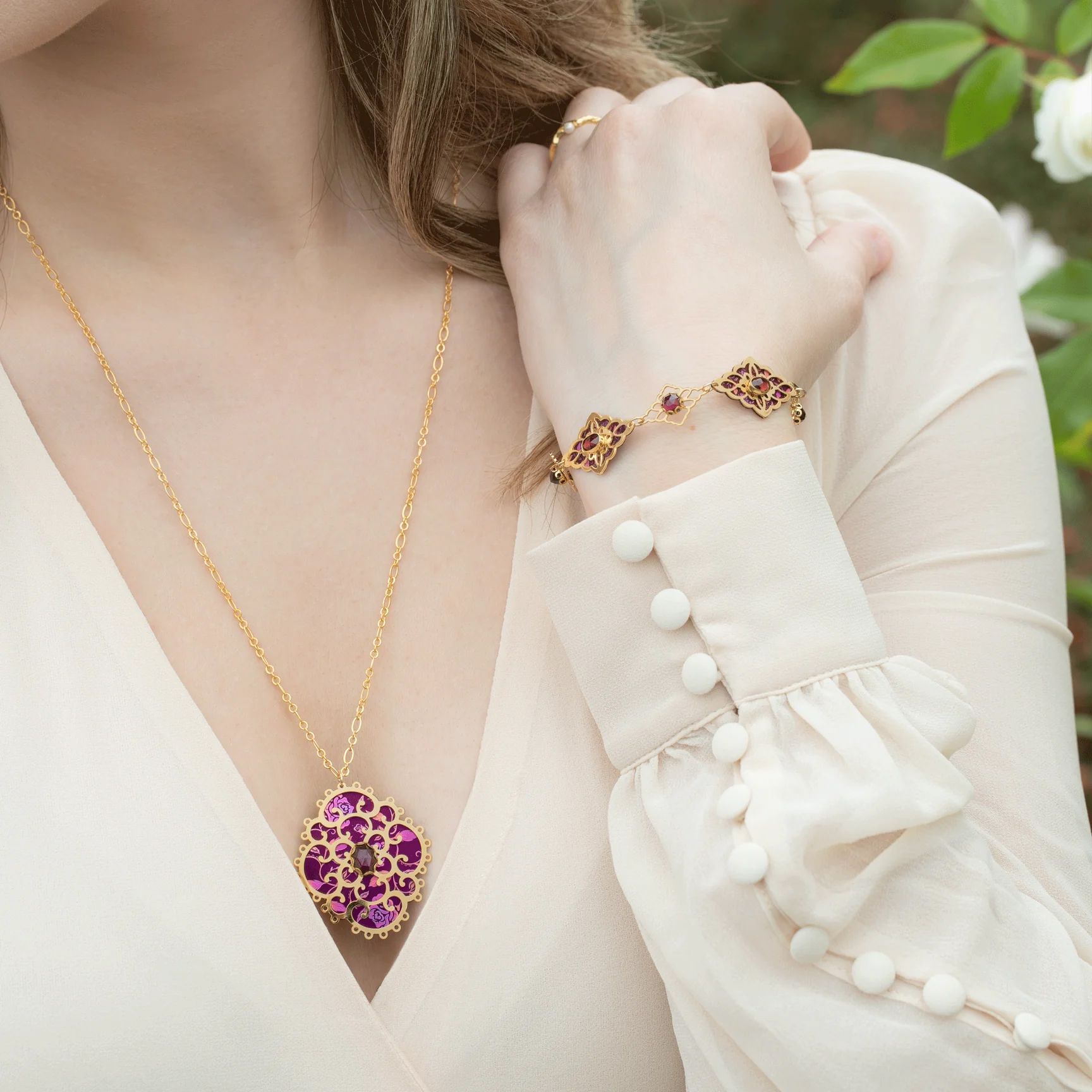 Woman wearing gold necklace and bracelet, with pink and purple gemstones.