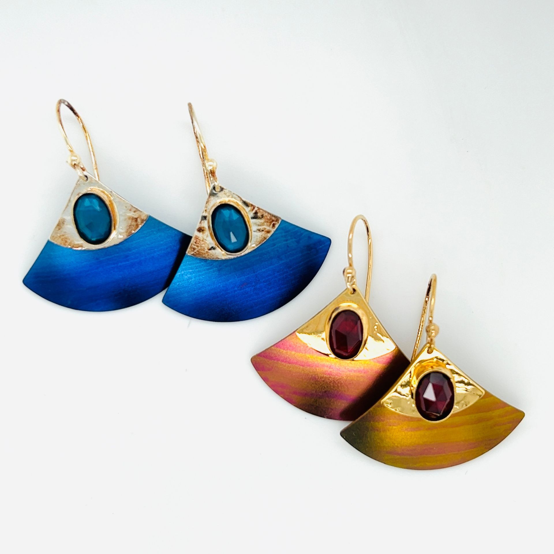 Two pairs of fan-shaped earrings. One pair is blue with a teal gem, the other is gold/purple with a red gem.