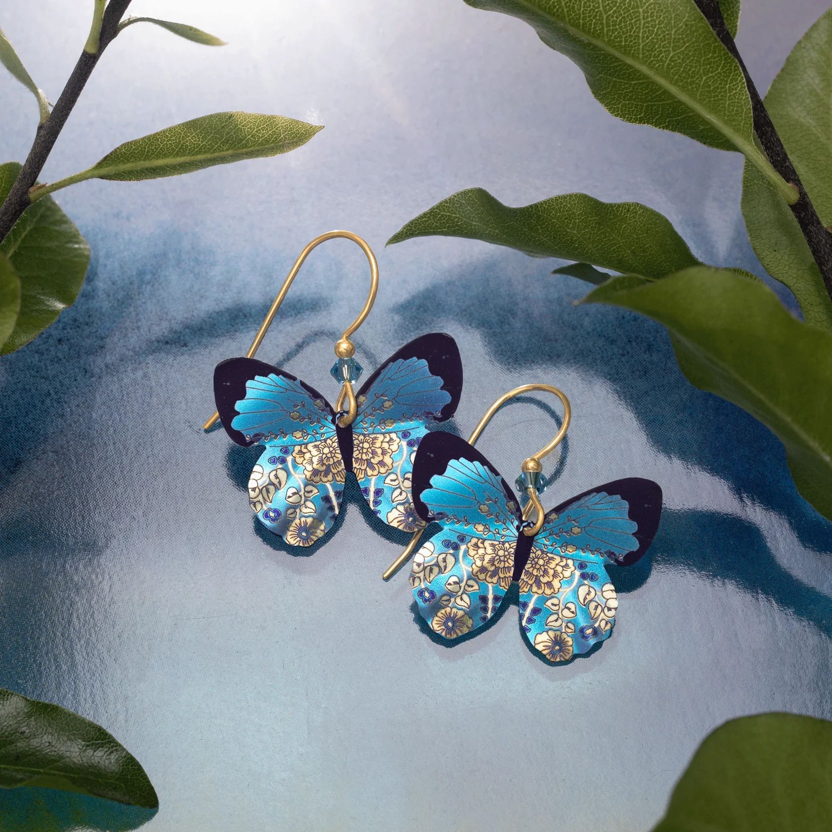 Pair of blue and gold butterfly earrings on a blue fabric background, with green leaves in the corners.