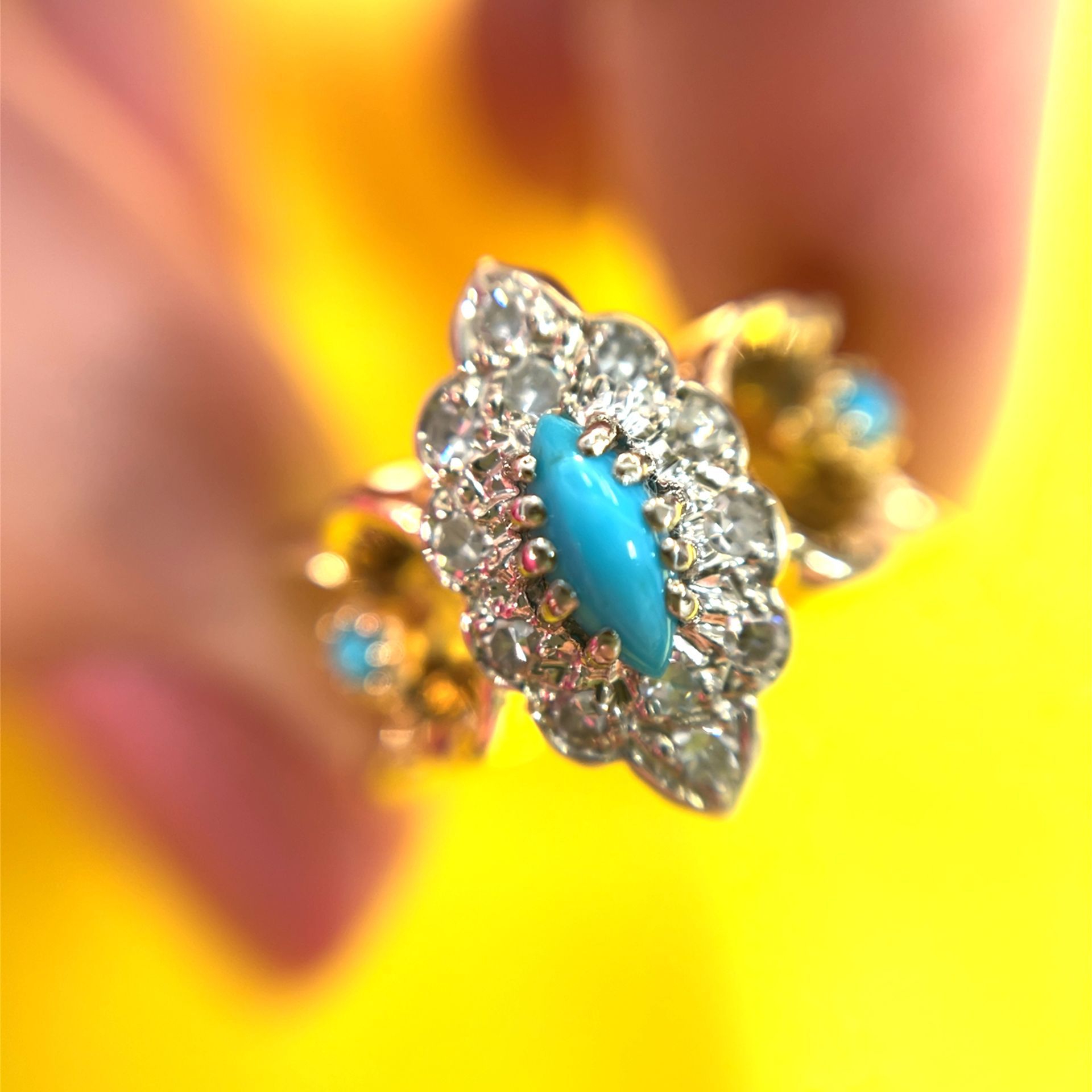 Close-up of a gold ring with a turquoise center stone surrounded by diamonds, held against a yellow background.