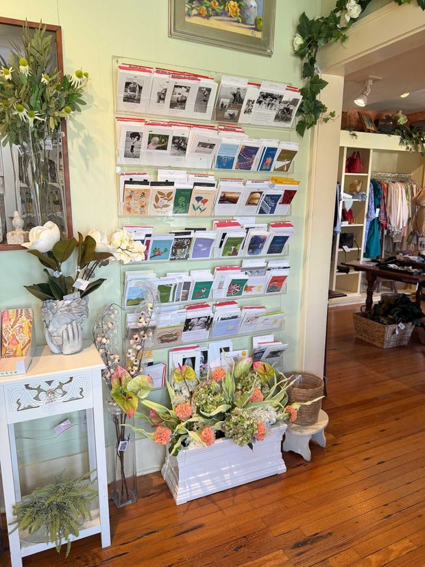A wall-mounted display of greeting cards in a shop, beside a floral arrangement on a white table and a wooden floor.