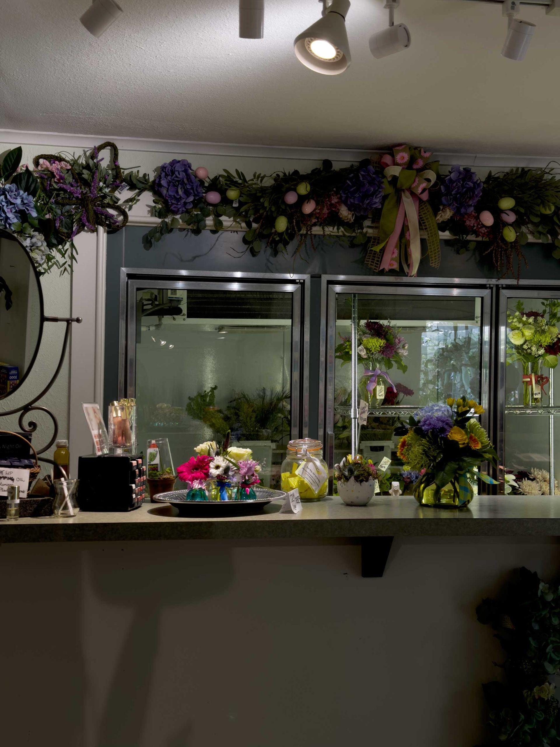 A floral shop counter with bouquets, colorful decorations, and glass display windows illuminated by ceiling spotlights.