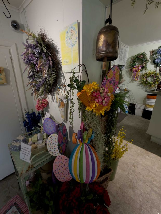 A display of colorful paper Easter eggs, floral arrangements, and wreaths inside a shop with a large hanging bell.