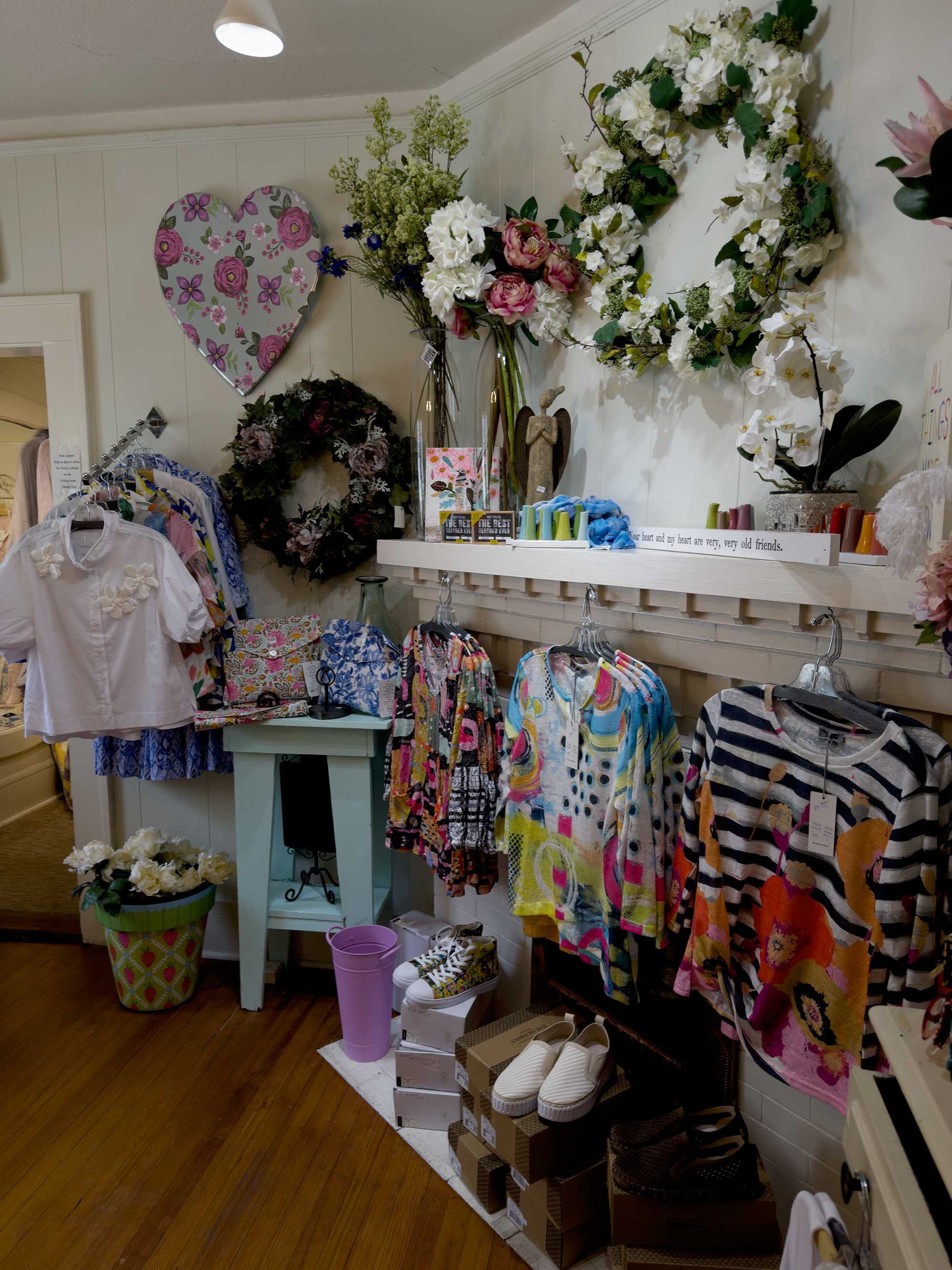 A boutique display features clothing racks with patterned tops, floral wreaths, a heart-shaped sign, and white shoes.