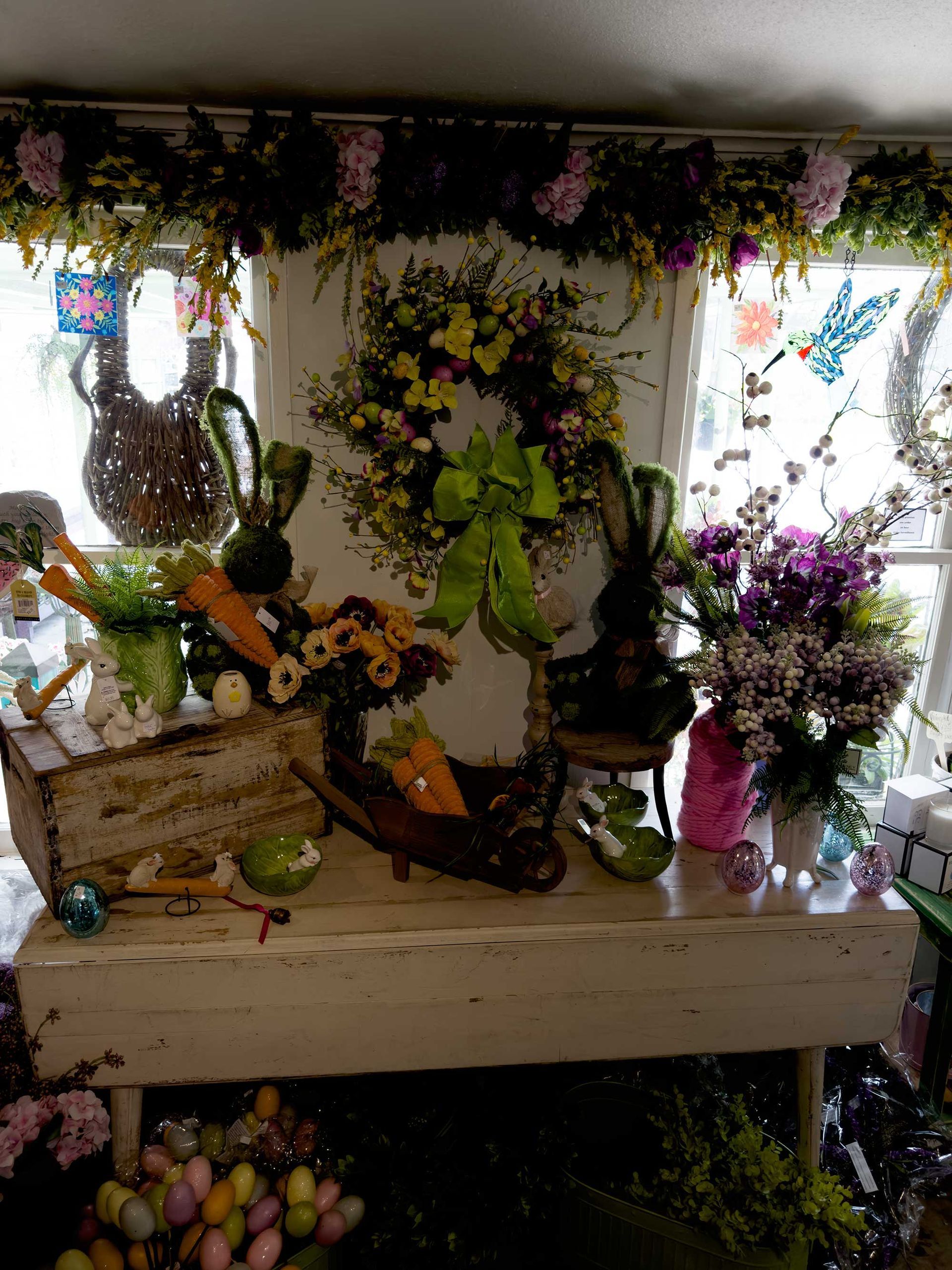 A floral Easter display on a white wooden table featuring a wreath, decorative bunnies, carrots, and a bowl of eggs.