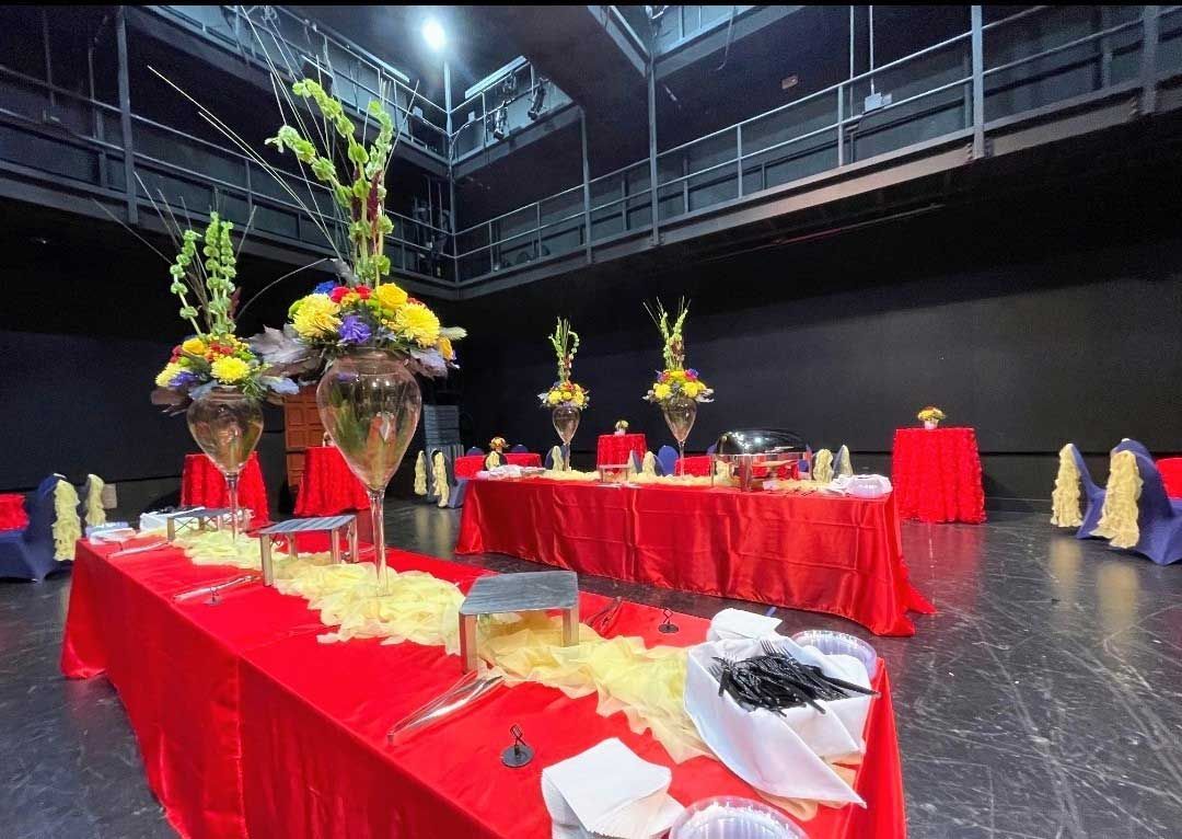 Two long banquet tables covered in red tablecloths are set for a formal event in a large, dark-walled event space.