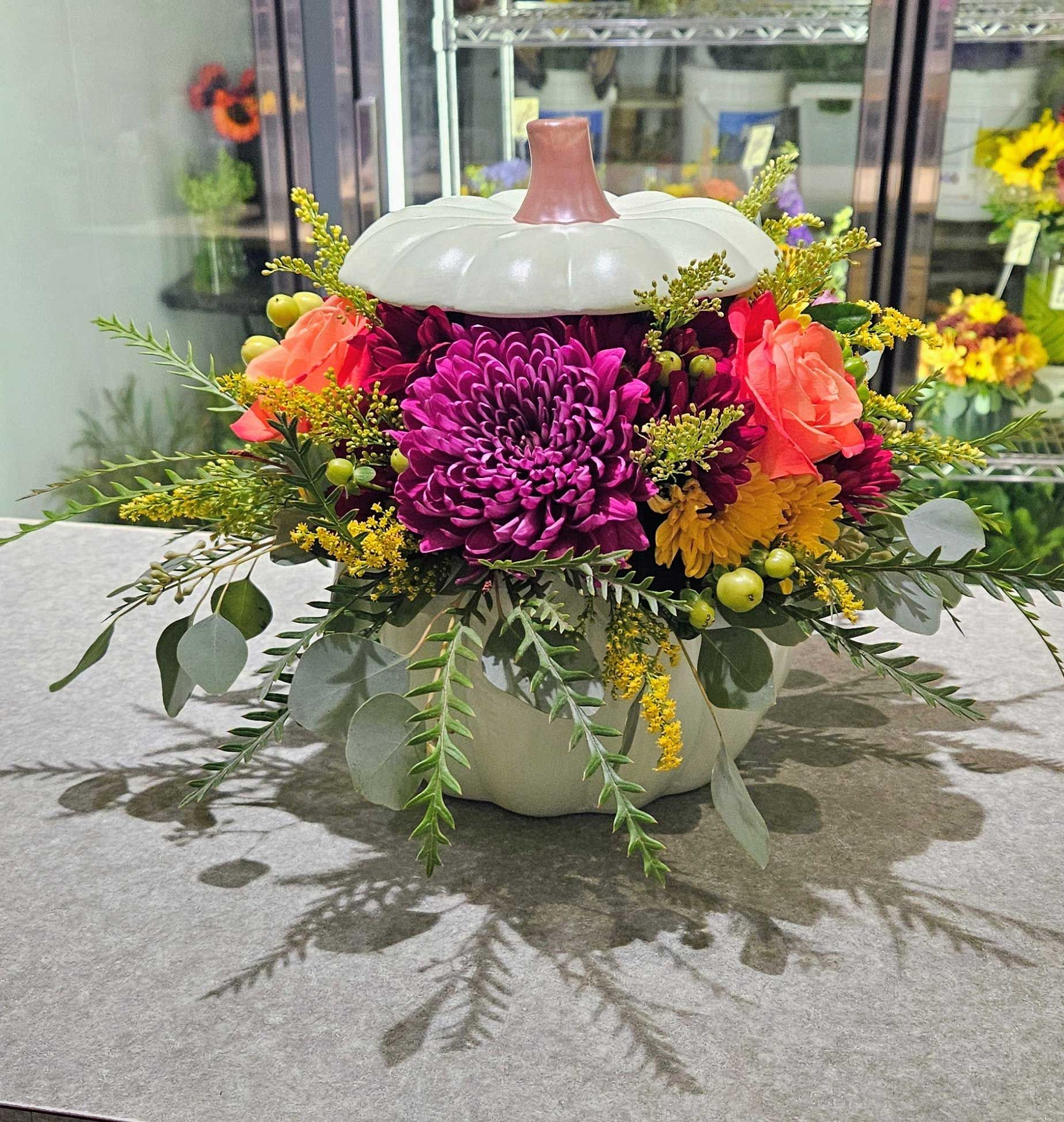 A festive pumpkin-shaped container holding a colorful floral arrangement of dark purple mums, orange roses, and greenery.