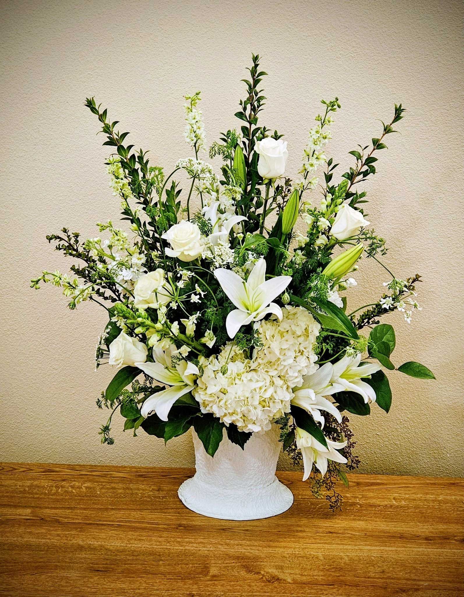 An arrangement of white lilies, roses, and hydrangeas with green foliage in a white vase on a wood table.