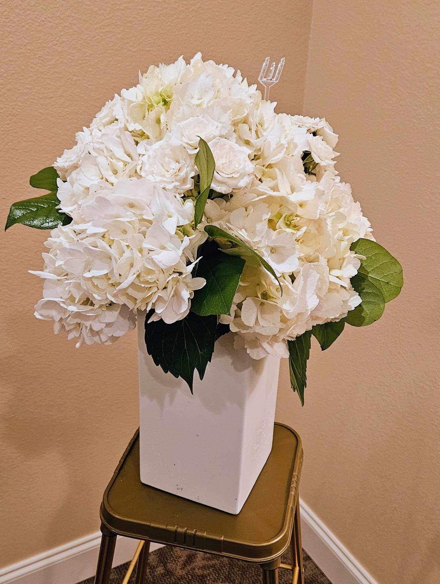 A large arrangement of white hydrangea flowers in a square white vase, resting on a small, gold-edged pedestal table.