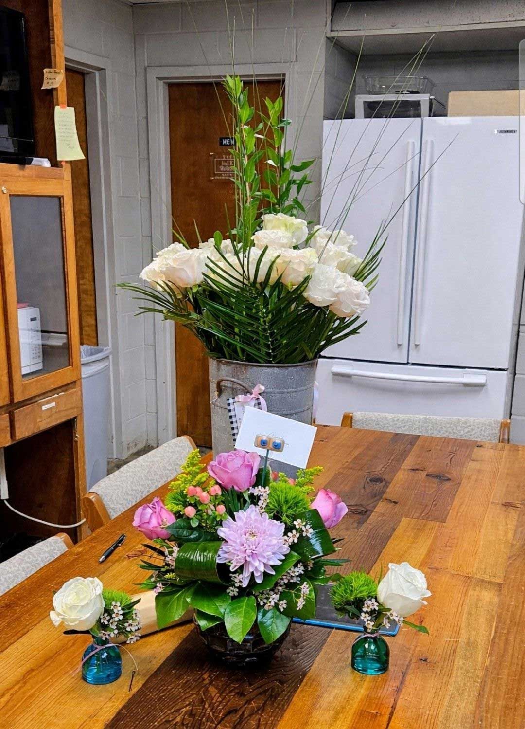 A tall white floral arrangement in a metal bucket sits behind a smaller centerpiece of pink and white flowers on a table.