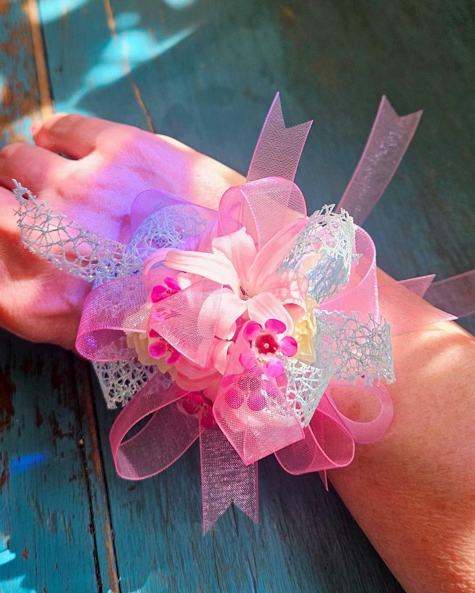 A decorative wrist corsage with pink fabric flowers and silver mesh ribbons, worn on a hand against a blue wooden surface.