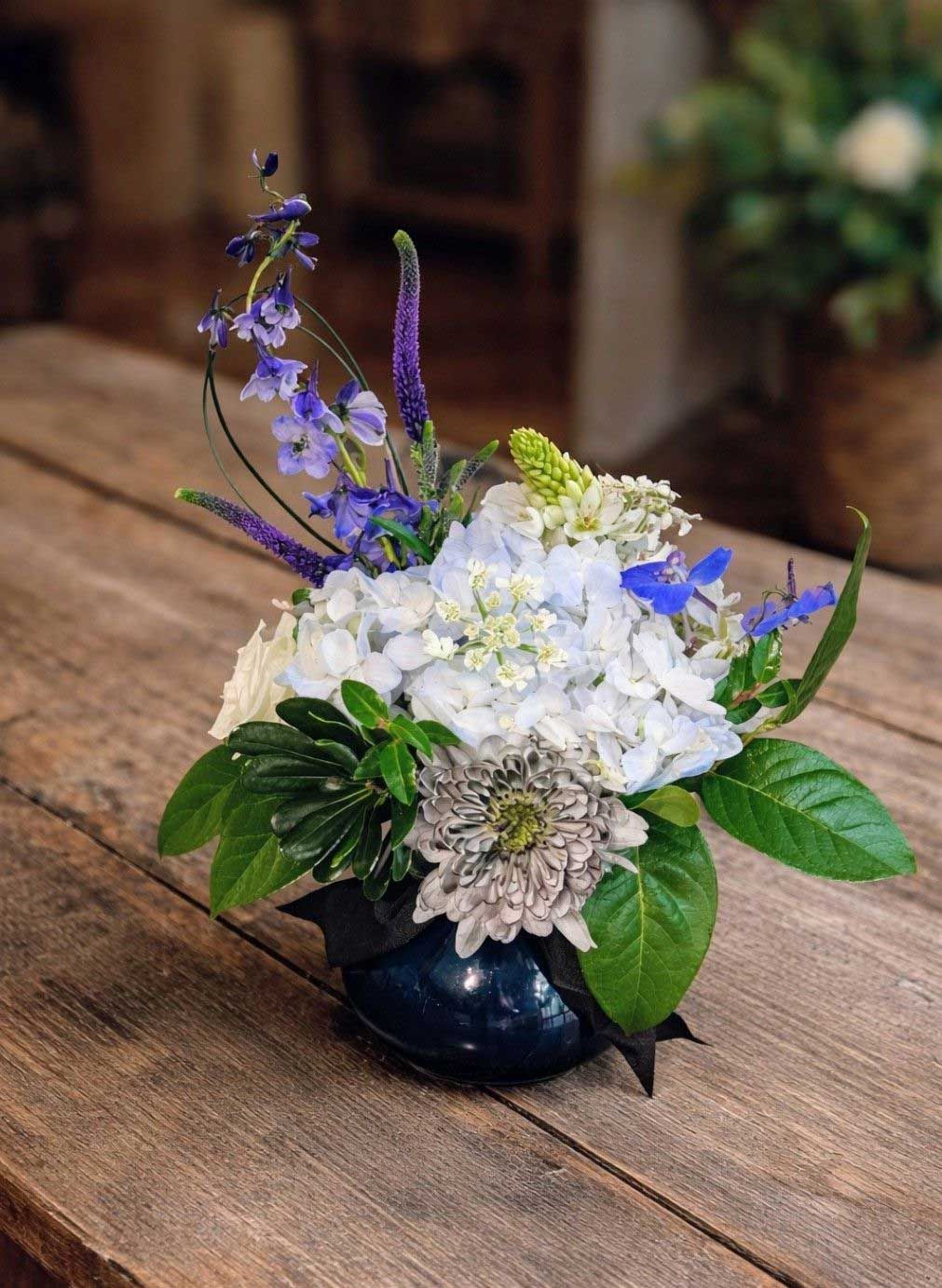 A blue ceramic vase holds a floral arrangement of blue hydrangeas, purple delphiniums, and white flowers on wood.