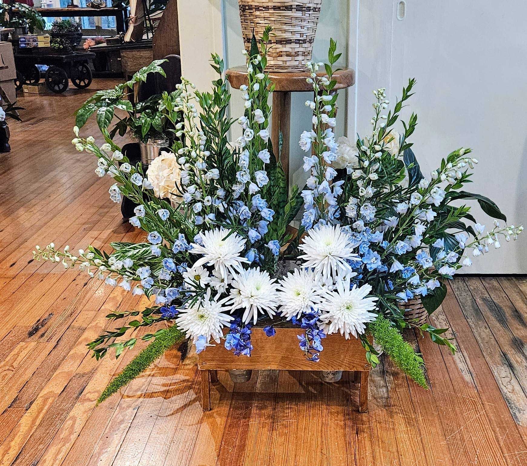 A floral arrangement of blue larkspur and white spider mums in a wooden crate, sitting on a wooden floor.