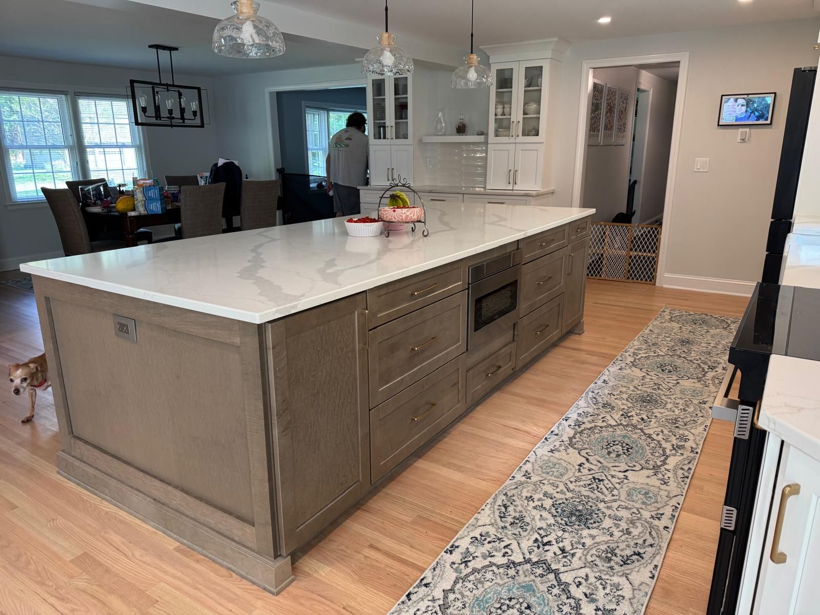 Kitchen with large island and white countertop. A person is visible in the background. Wooden floors and patterned rug.