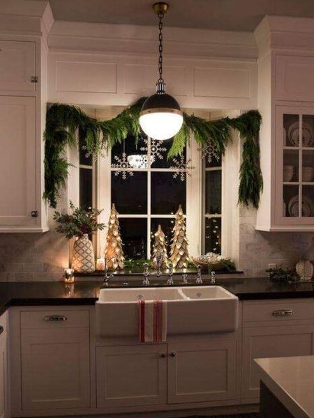 Kitchen window decorated for Christmas, featuring garland, snowflakes, and illuminated trees.