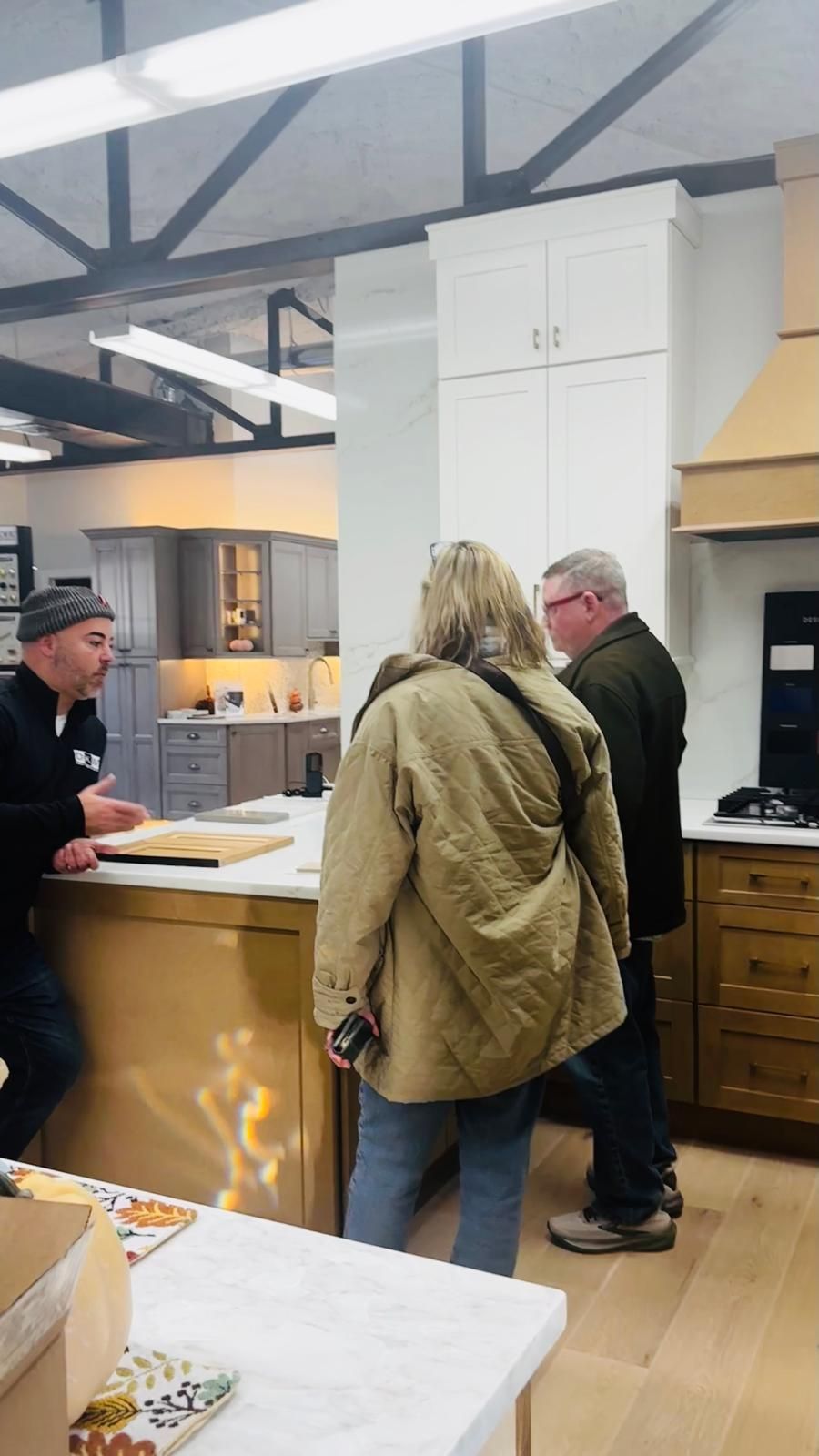 A person in a black shirt speaks to a couple near a kitchen display; the setting is a showroom.