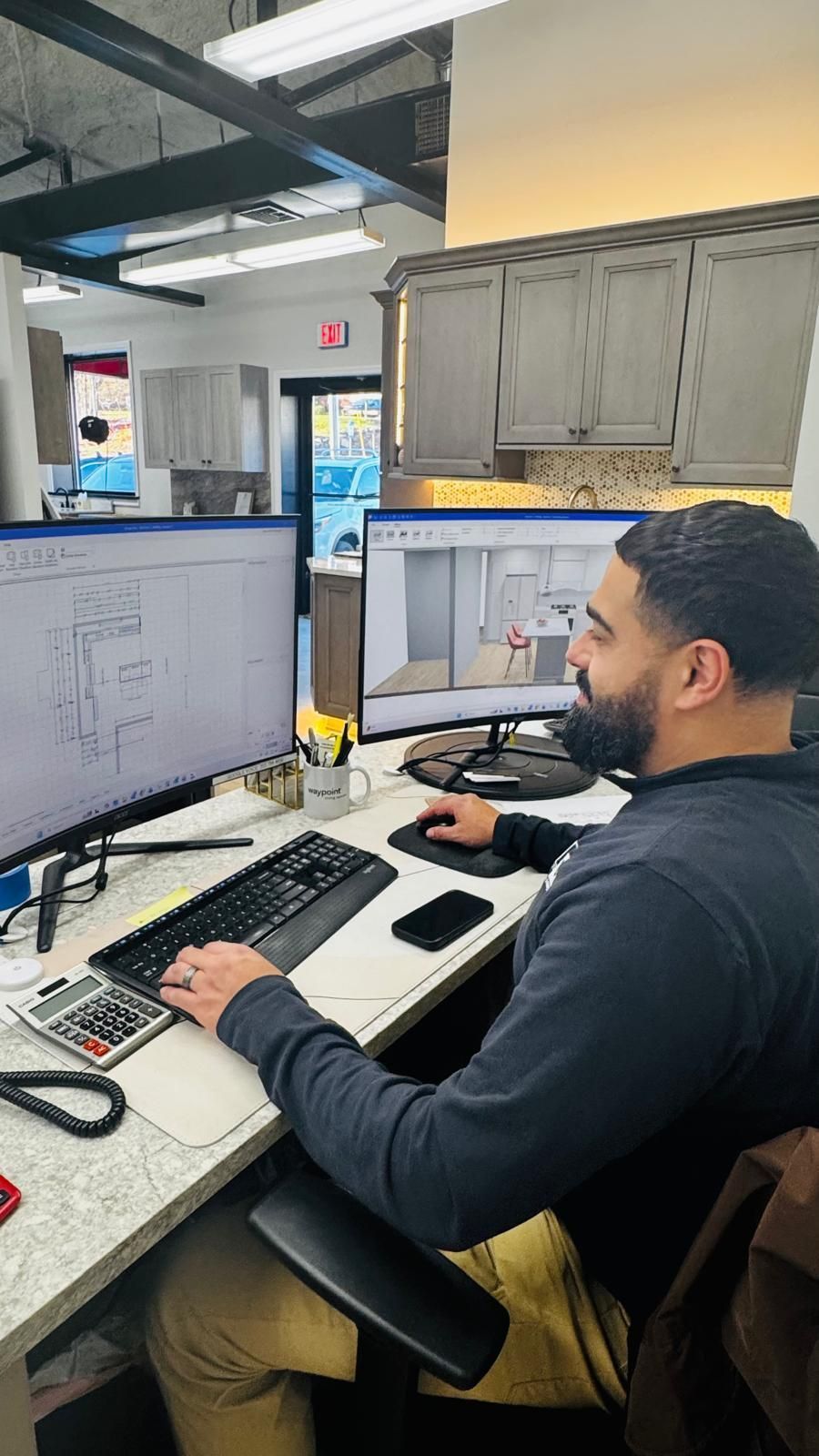 A man at a desk is working on two computer screens with design plans. Office setting, focused expression.