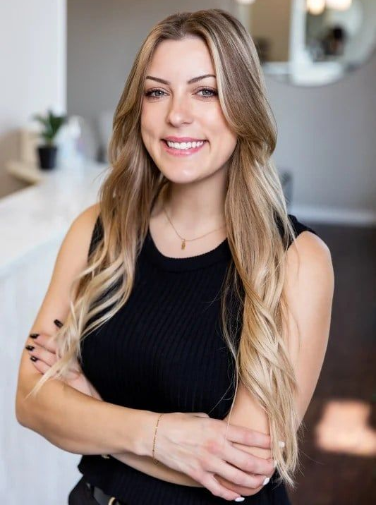 Woman with blonde hair smiling, arms crossed. Wearing a black top in a bright interior.