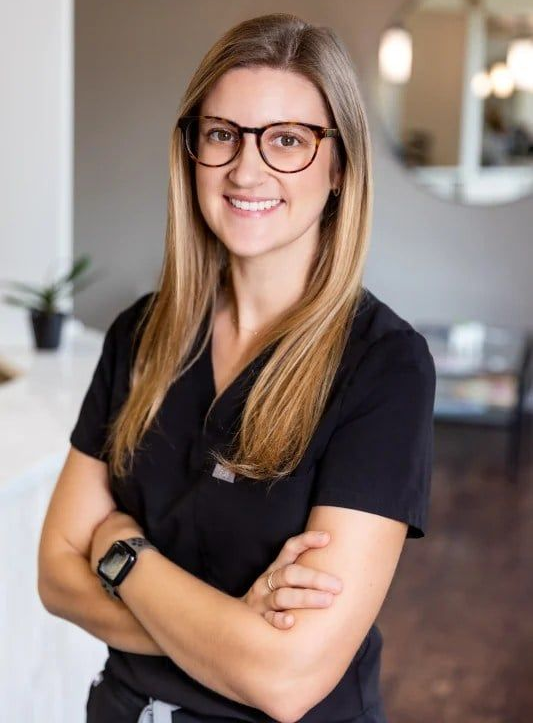 Woman in black scrubs and glasses, arms crossed, smiling in a modern office.