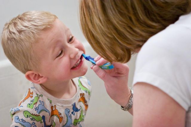 A person brushing a child's teeth in a bathroom; both are smiling.