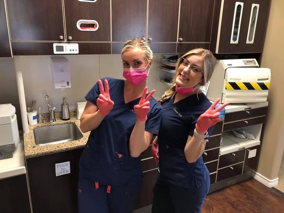 Two dental assistants in blue scrubs and pink masks give peace signs in a dental office.