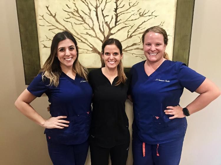Three people posing. Center person in black, flanked by two in blue scrubs, arms akimbo, smiling. Artwork backdrop.