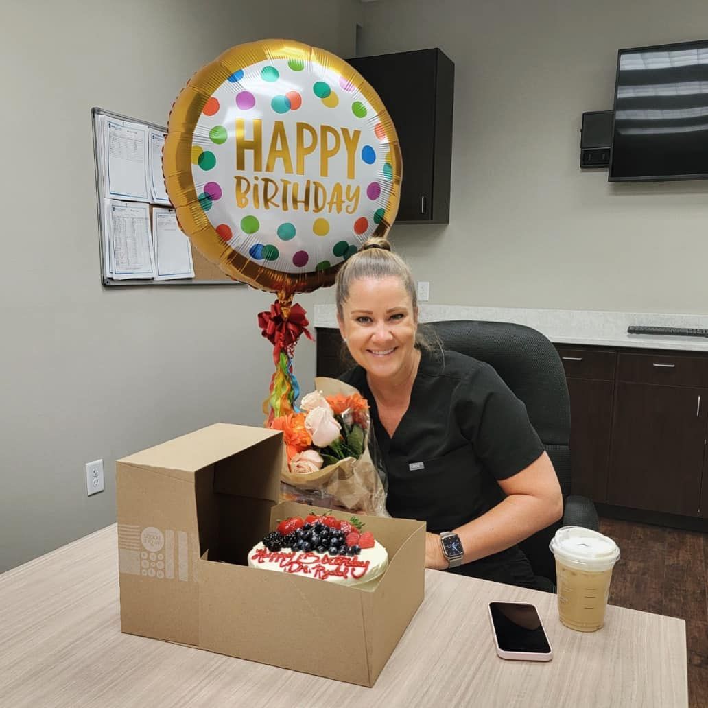 Woman in scrubs smiles at desk with birthday balloon, cake, flowers, and coffee.