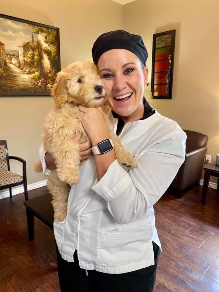 Woman in white coat and black head covering holding a tan Goldendoodle puppy, smiling.