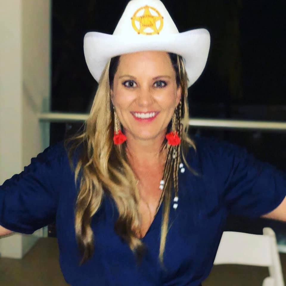 Woman wearing a cowboy hat and dark blue shirt, smiling, with red earrings.
