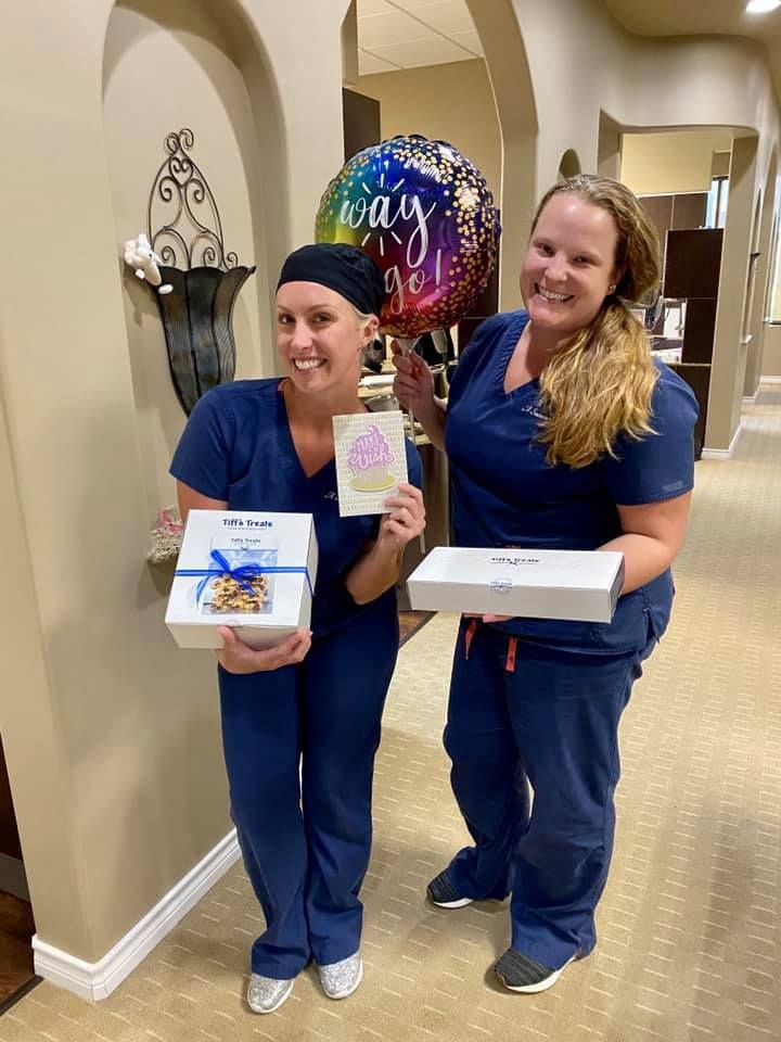 Two women in scrubs smile, holding gifts, cards, and a 