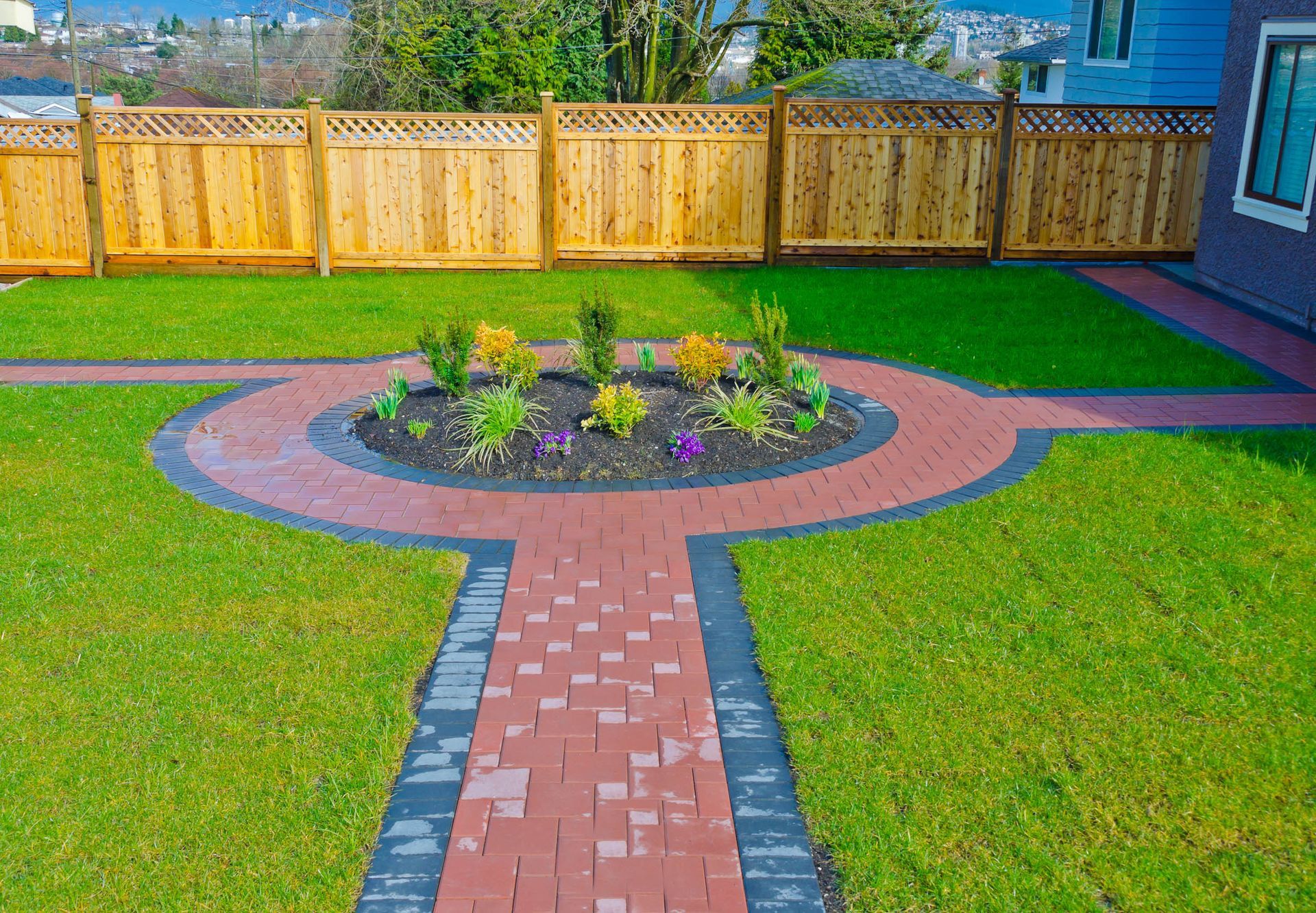 Backyard garden with brick path, circular flower bed, green lawn, and wooden fence