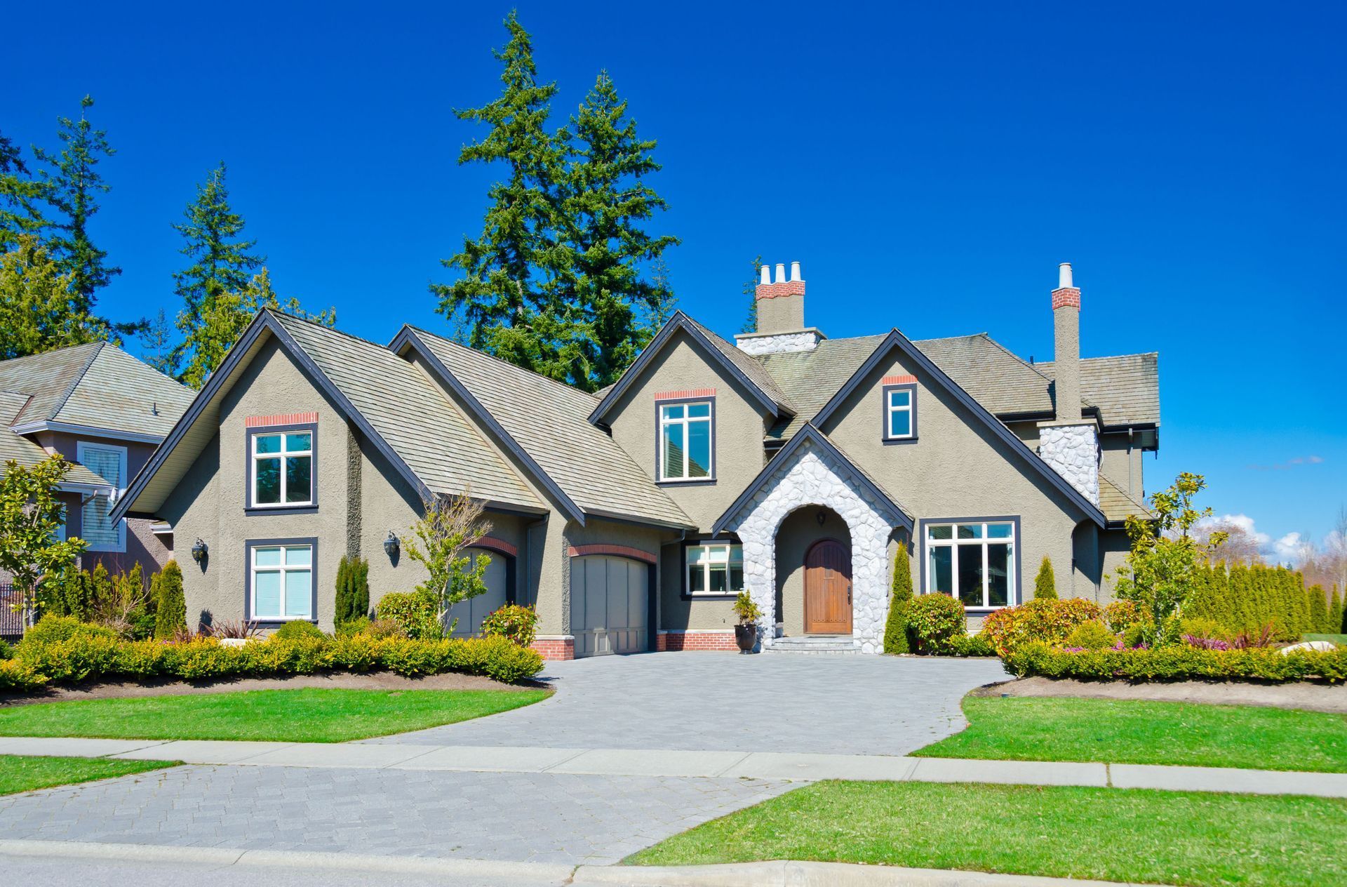 Large suburban house with gray roof, arched stone entry, and landscaped lawn under a clear blue sky