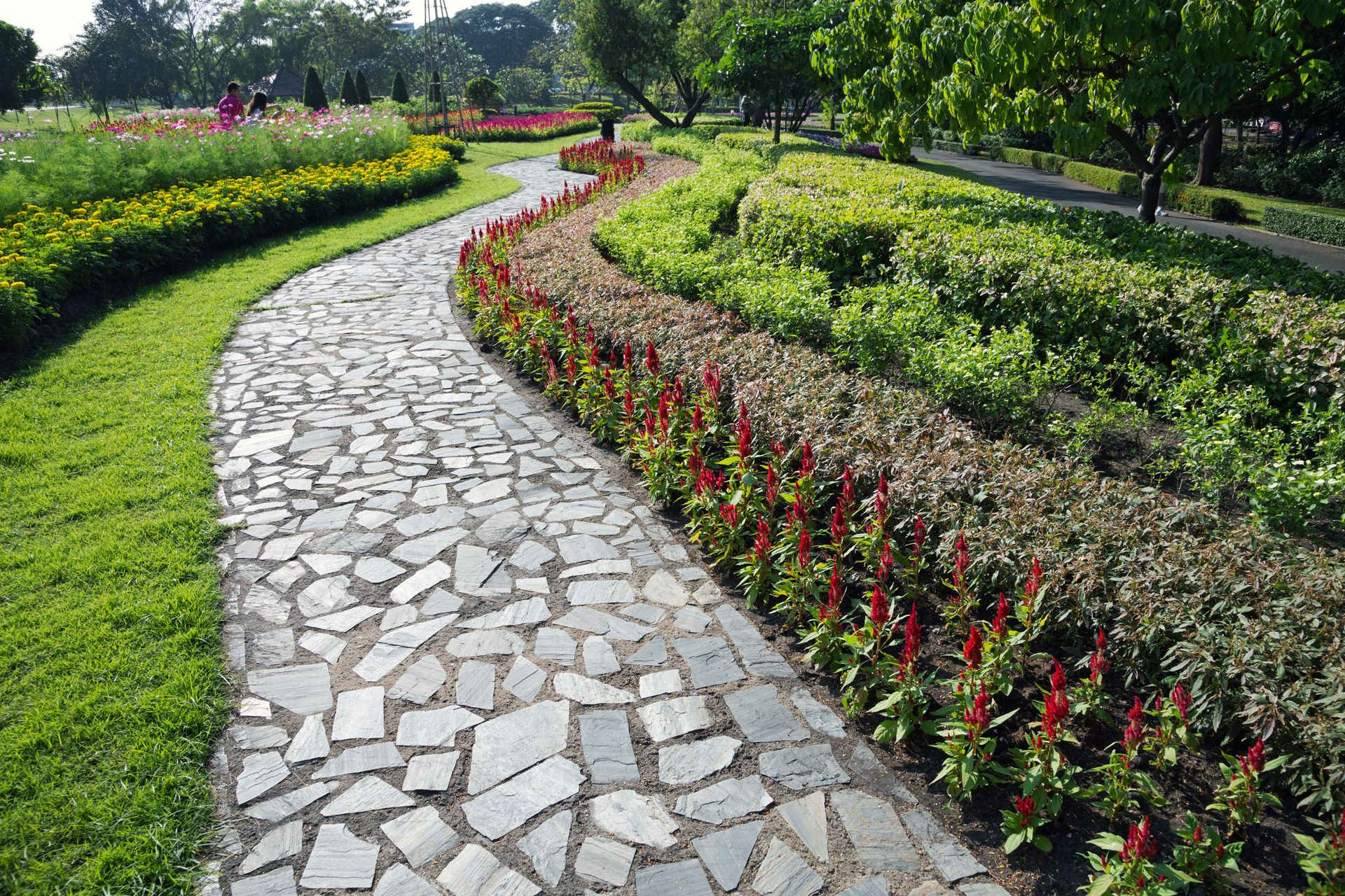 Curved stone garden path bordered by red flowers and green hedges under trees