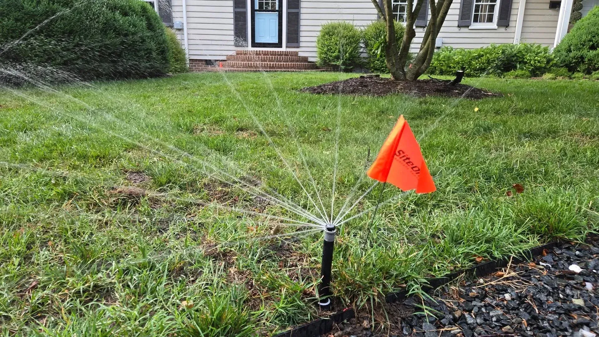Lawn sprinkler spraying water on a green yard, with an orange marker flag nearby.