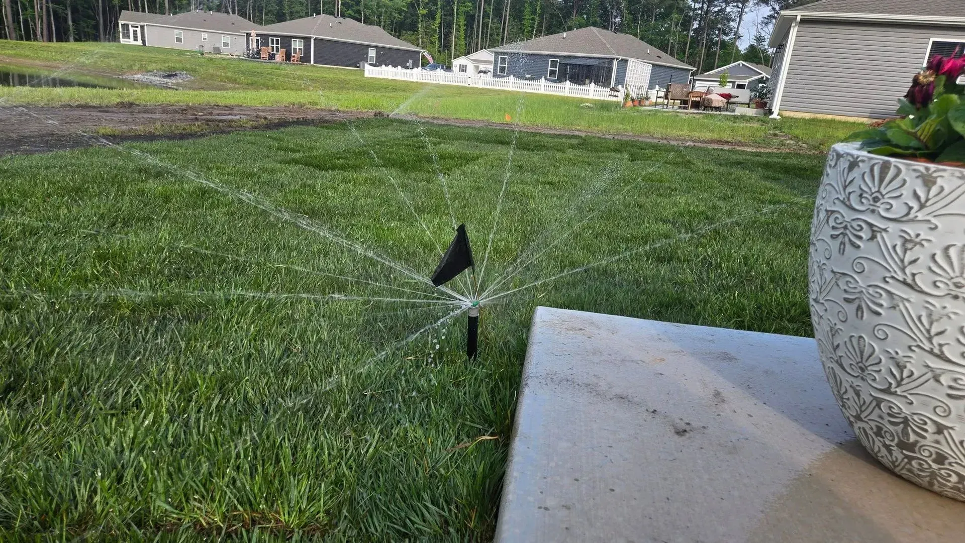 Sprinkler spraying water across a green lawn beside a patio and flower pot.