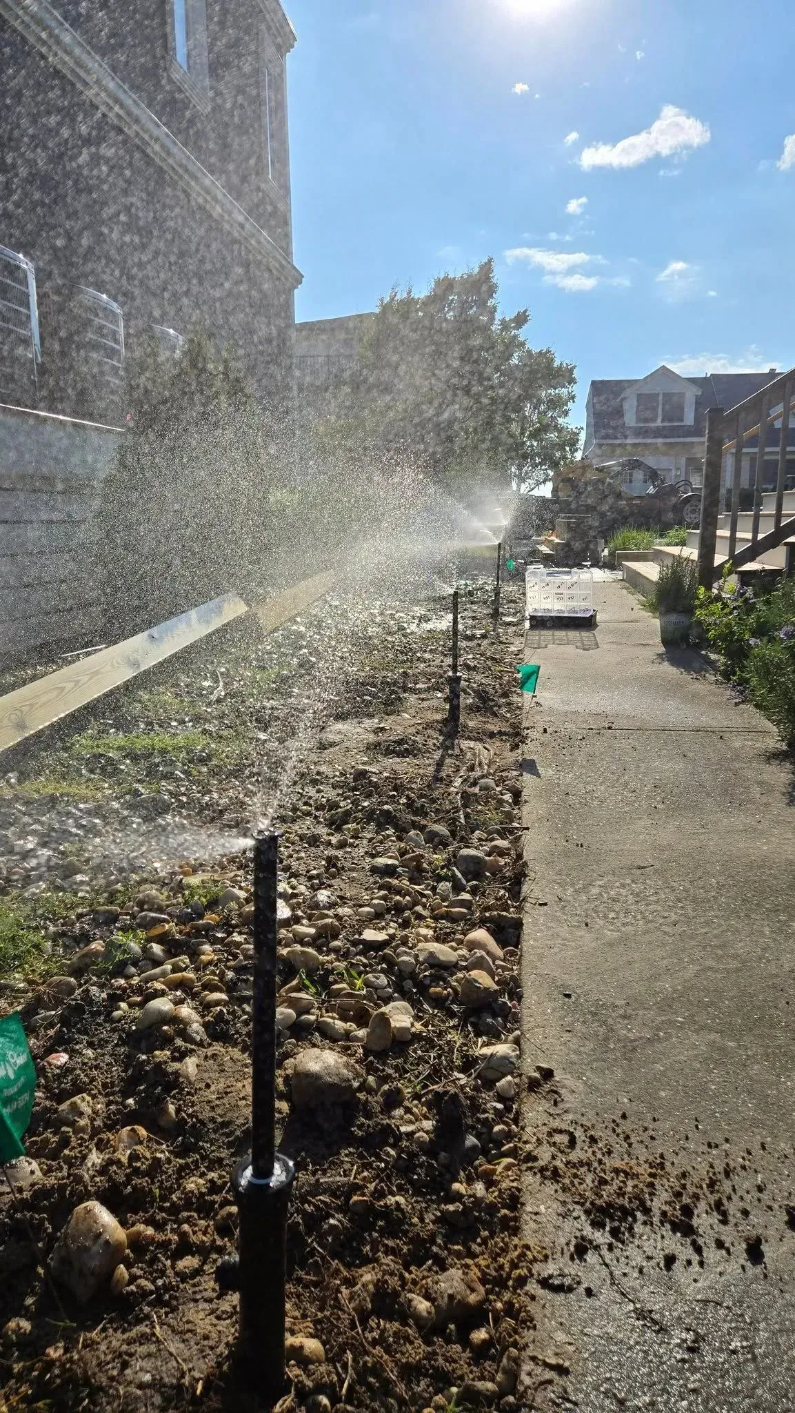 Sunlit walkway beside a stone wall with a sprinkler spraying water over newly laid gravel and a residential building ahead
