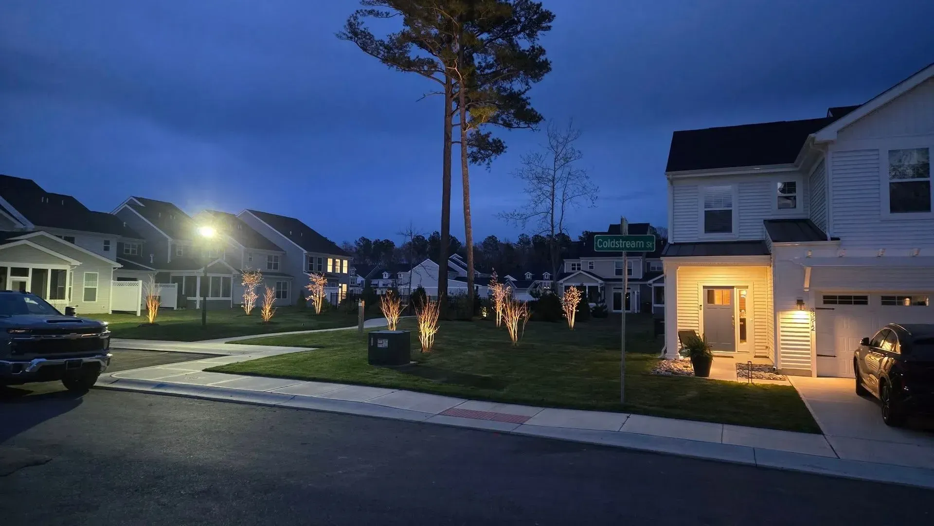 Suburban street at dusk with lit houses, parked cars, and a grassy median with young trees.