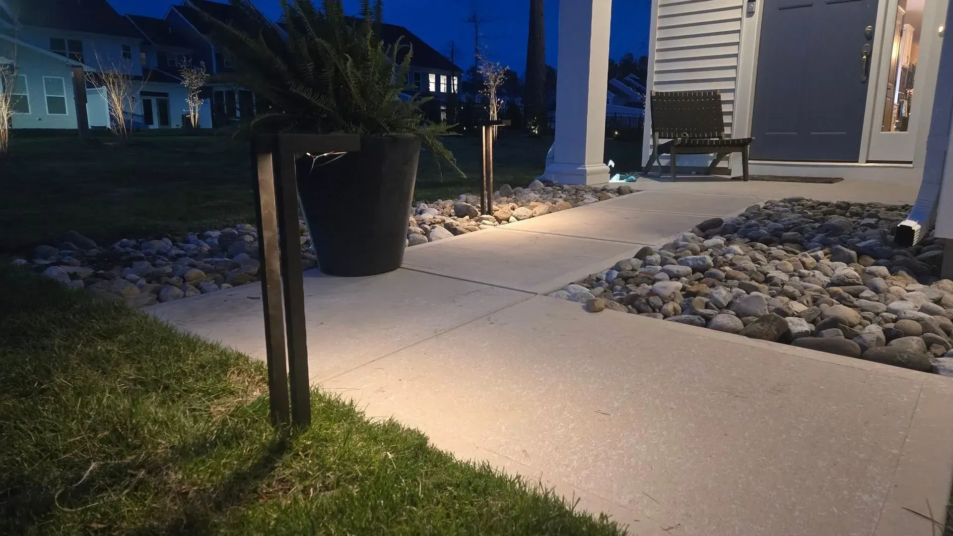 Lit front walkway with a black planter, rocks, and a house entrance at dusk