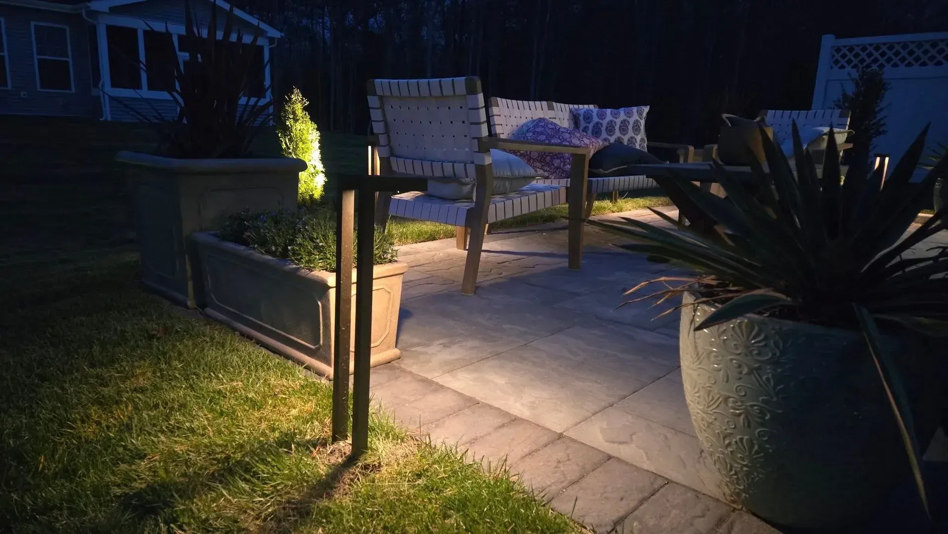 Nighttime patio with two chairs and a small table on a tiled path, lit by outdoor lights.